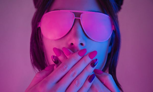 Young woman in pink glasses covers her mouth with hands with a manicure, retrowave style portrait. Demonstration of a beautiful manicure.